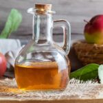 Glass bottle of apple cider vinegar in front of basket with apples