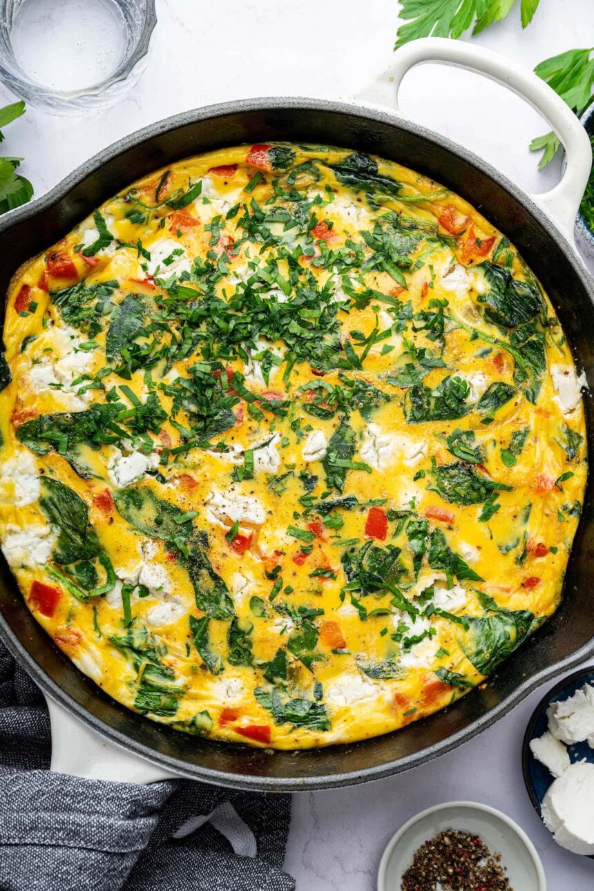 A cooked frittata in a white cast iron skillet topped with fresh parsley. There is a kitchen towel covering the skillet handle.