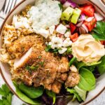 A bowl served with mixed greens, Greek chicken, tzatziki, hummus, cucumber tomato salad and feta cheese.