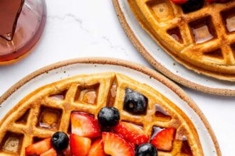 Golden brown protein waffles on a plate, topped with fresh strawberries and blueberries, drizzled with syrup, and served with a side of fruit.