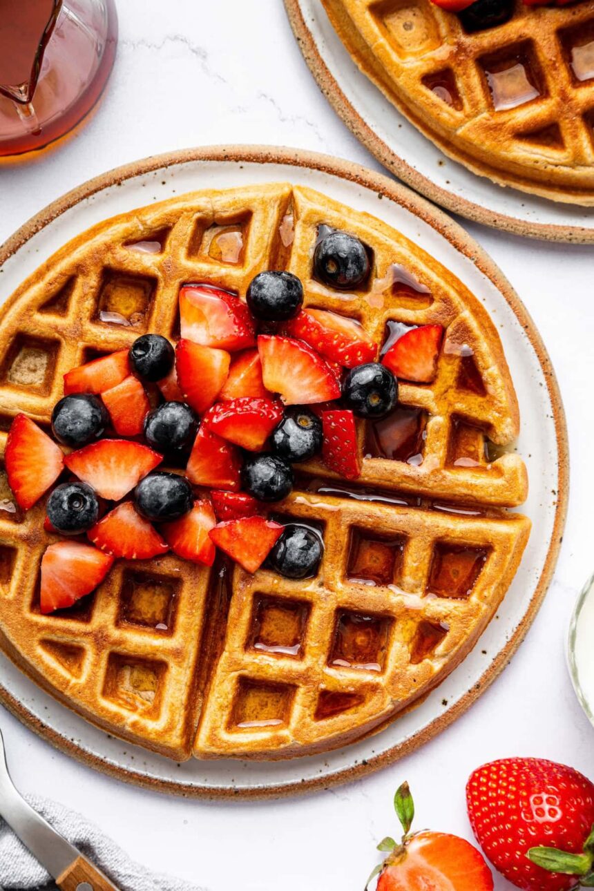 Golden brown protein waffles on a plate, topped with fresh strawberries and blueberries, drizzled with syrup, and served with a side of fruit.