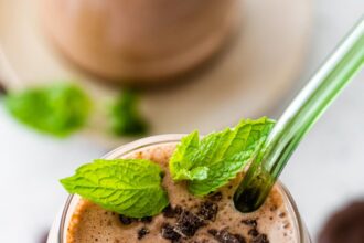 One mint chocolate protein shake in a glass topped with crushed Thin Mint cookies and fresh mint. There is a green straw in the glass.