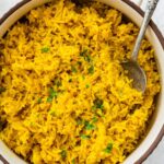 A pot of cooked turmeric rice topped with fresh parsley. There is a silver spoon in the rice.